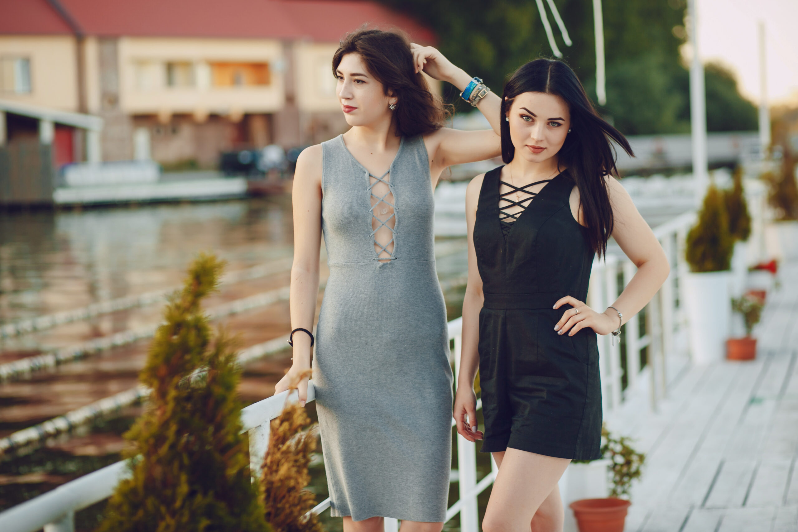 Two stylish girls in gorgeous dresses stand on the pier near the water