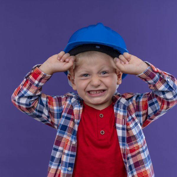 a cute little boy with blonde hair wearing checked shirt in blue helmet raising clenched fists looking at camera on a purple background