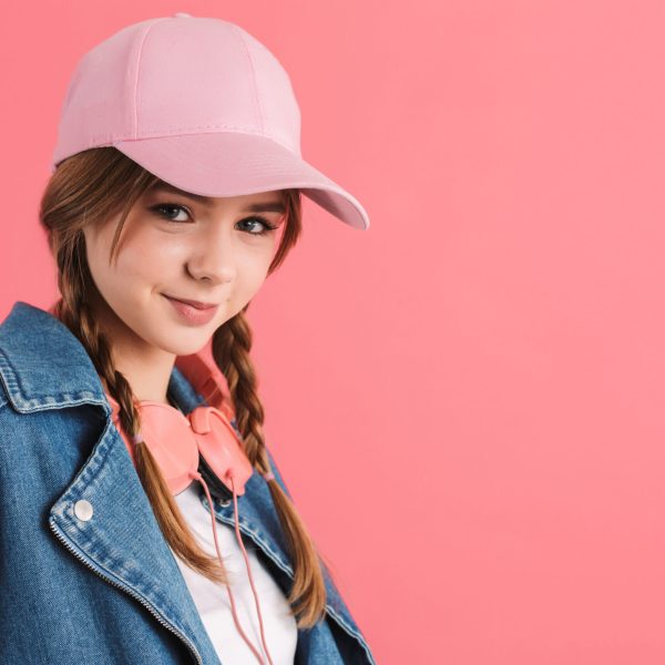 Portrait of young pretty joyful girl with two braids in denim jacket and cap with headphones on neck happily looking in camera over pink background